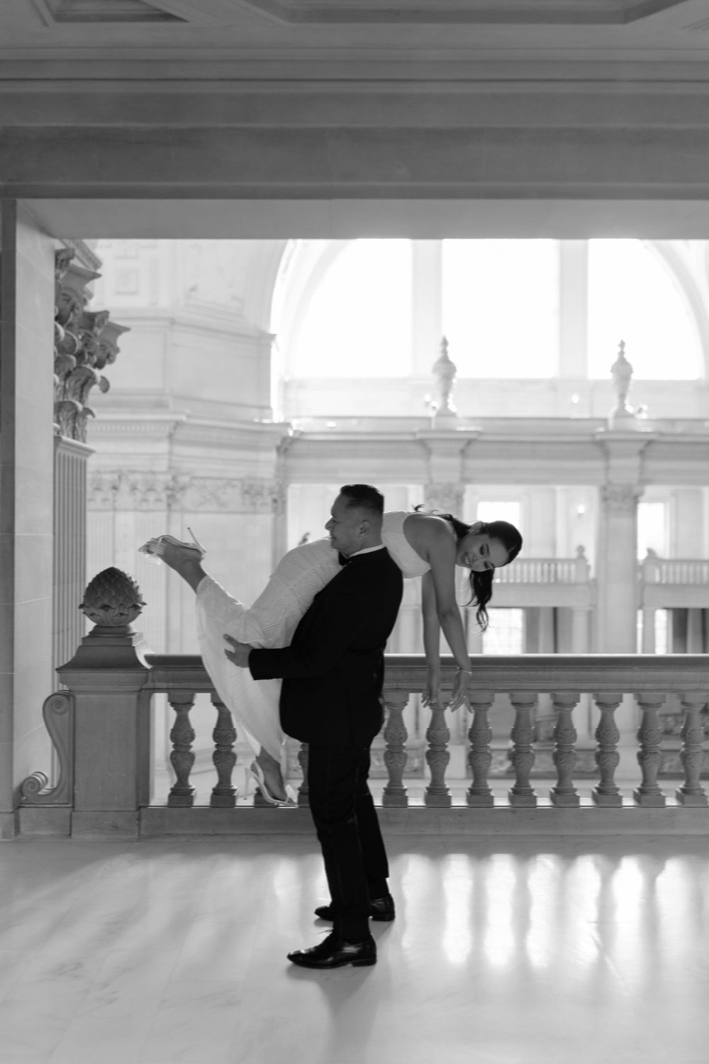 Black and white romantic ballroom dance pose with elegant couple in a grand white columned interior.