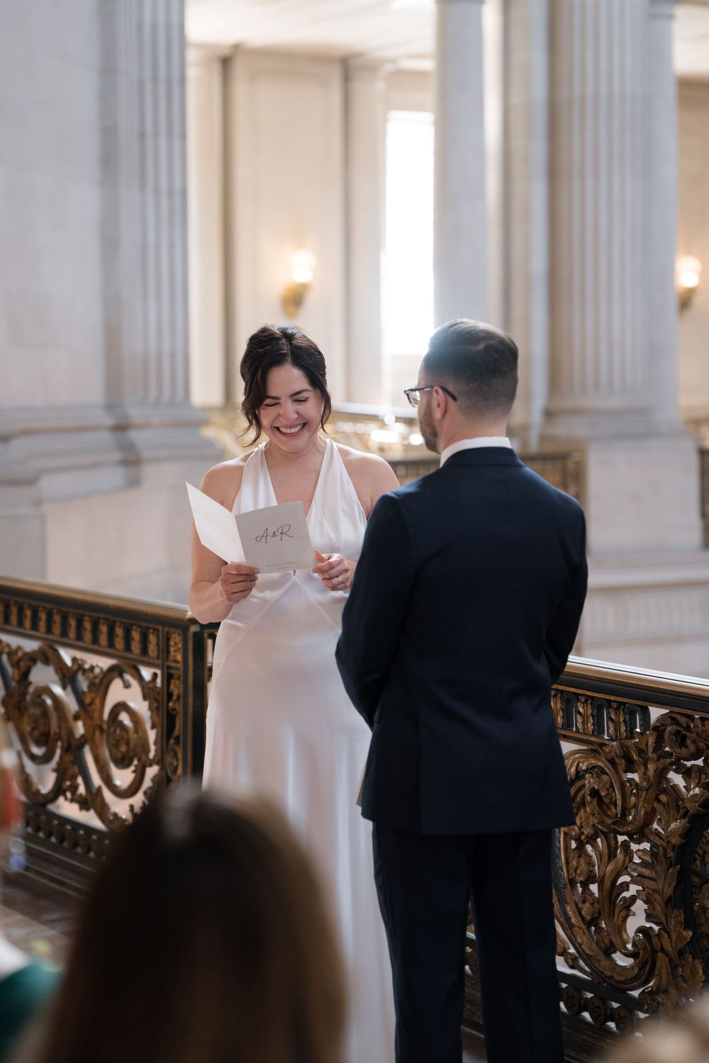 Wedding ceremony at elegant venue with ornate balcony railing and marble columns glowing with warm lighting.