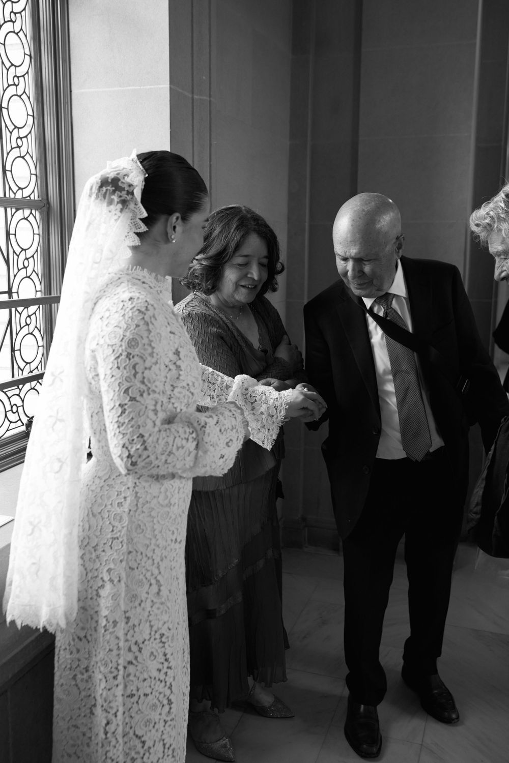 Black and white candid photo of people gathered near a window, with a bride in a delicate lace wedding dress.