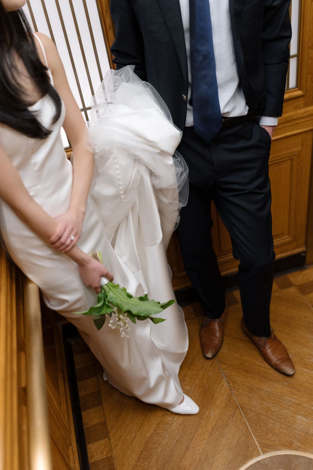 Close-up view of a white wedding dress and groom's suit, with the bride holding delicate green flowers on hardwood flooring.