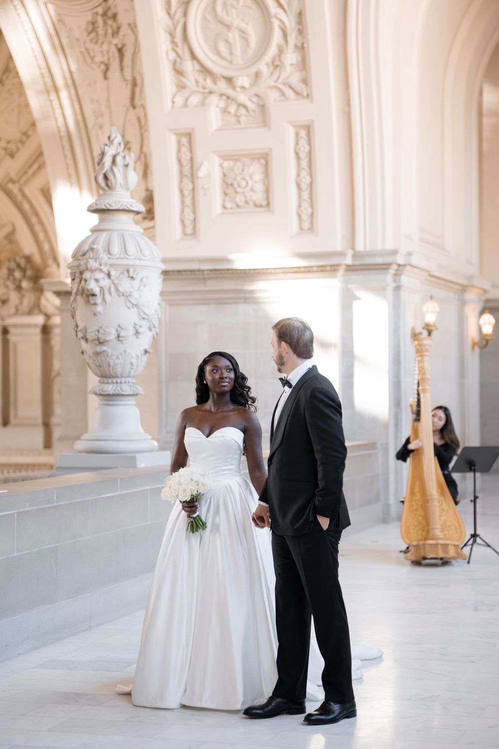 A couple in wedding attire stands in an elegant white marble hallway with ornate decor and a harpist in the background.