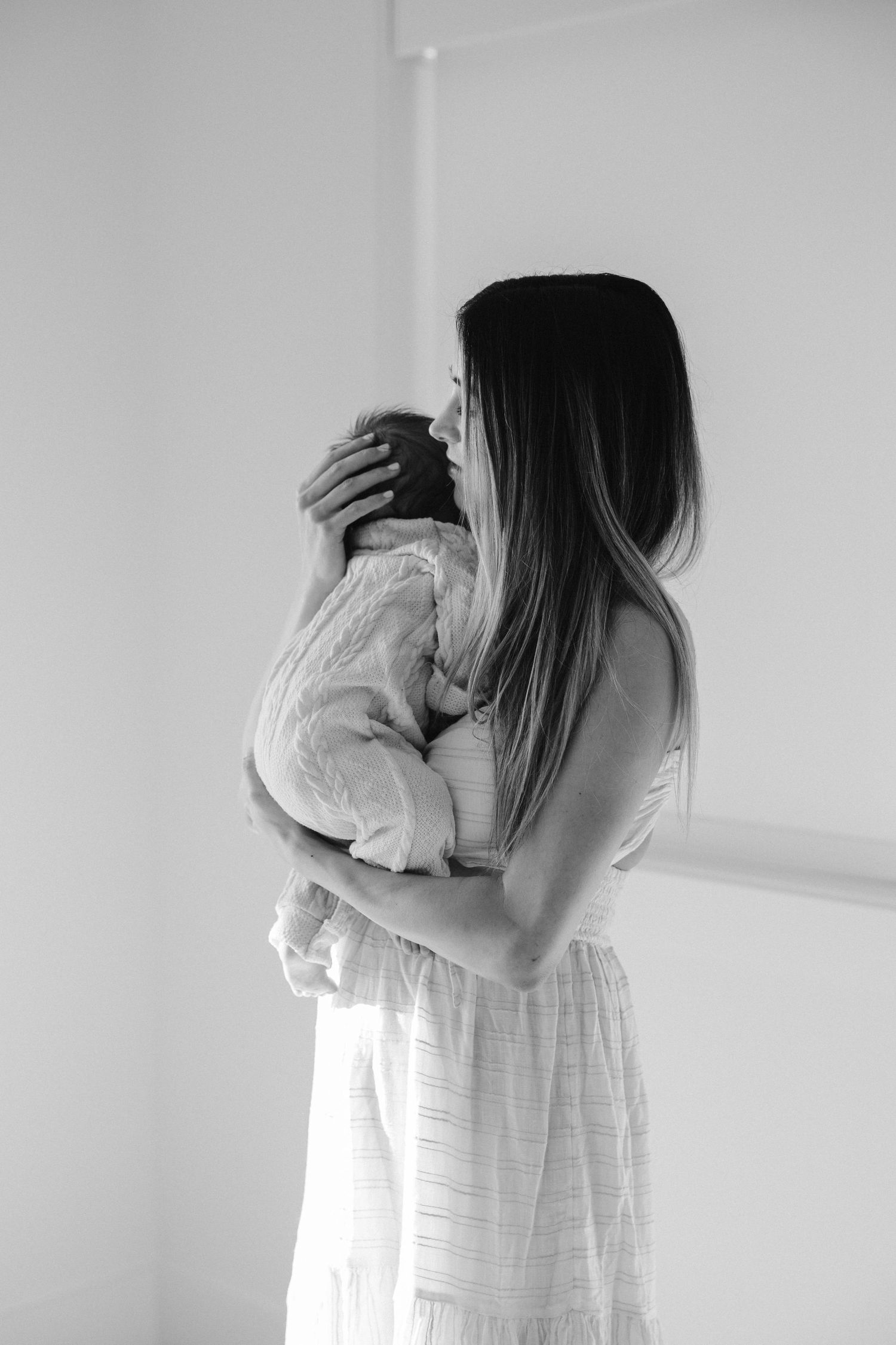 A woman in a white dress holds a baby against her chest in a minimalist black and white photograph.