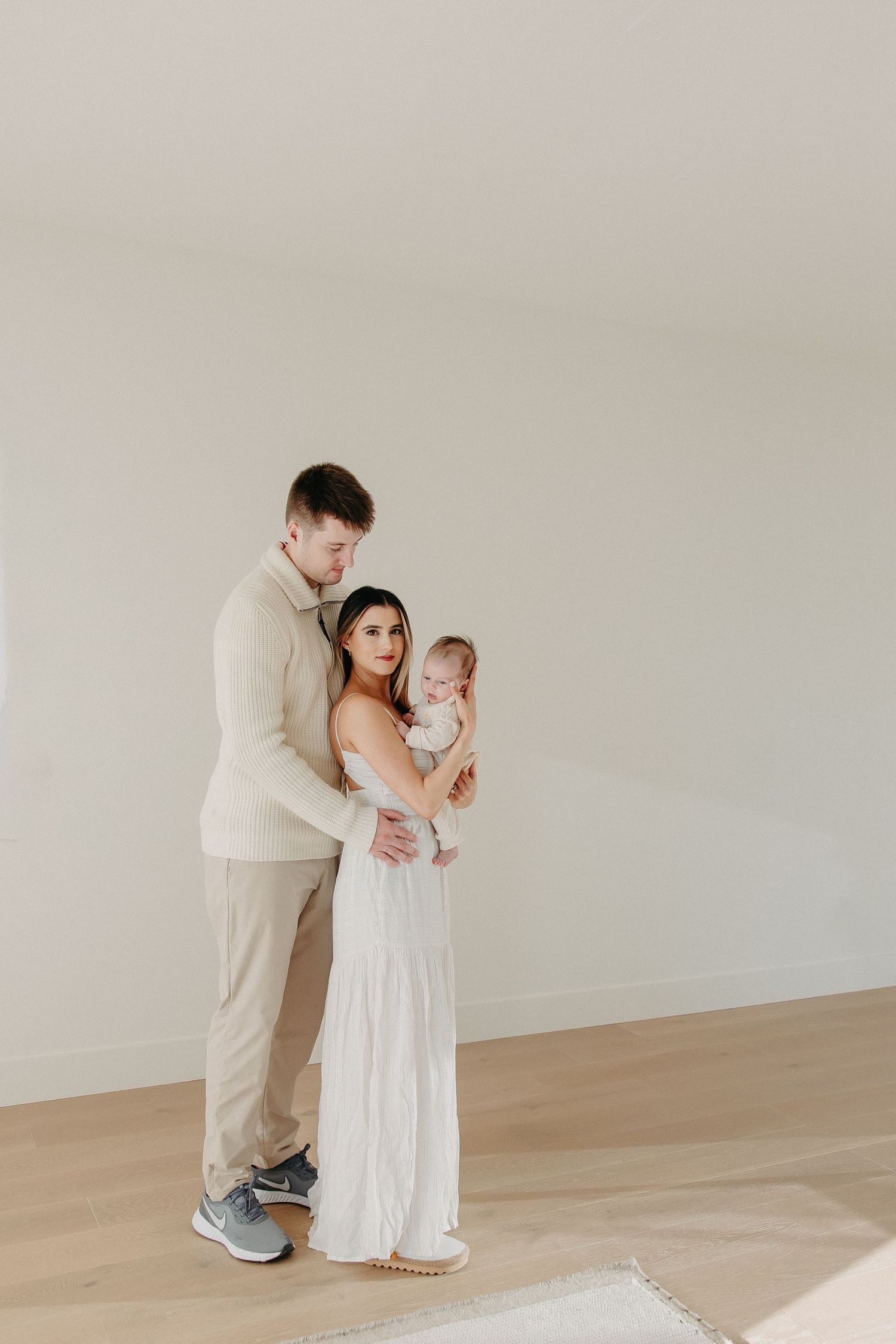 A couple in light-colored clothing stands together holding their baby in a minimalist room with wooden floors.