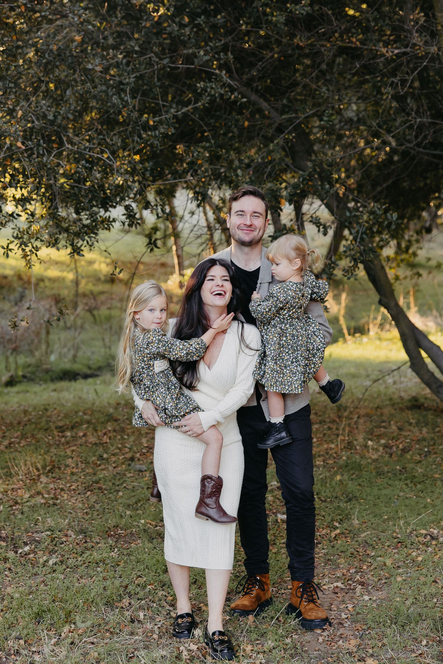 A family of four poses together outdoors under a tree during a sunny autumn day, wearing coordinating outfits.