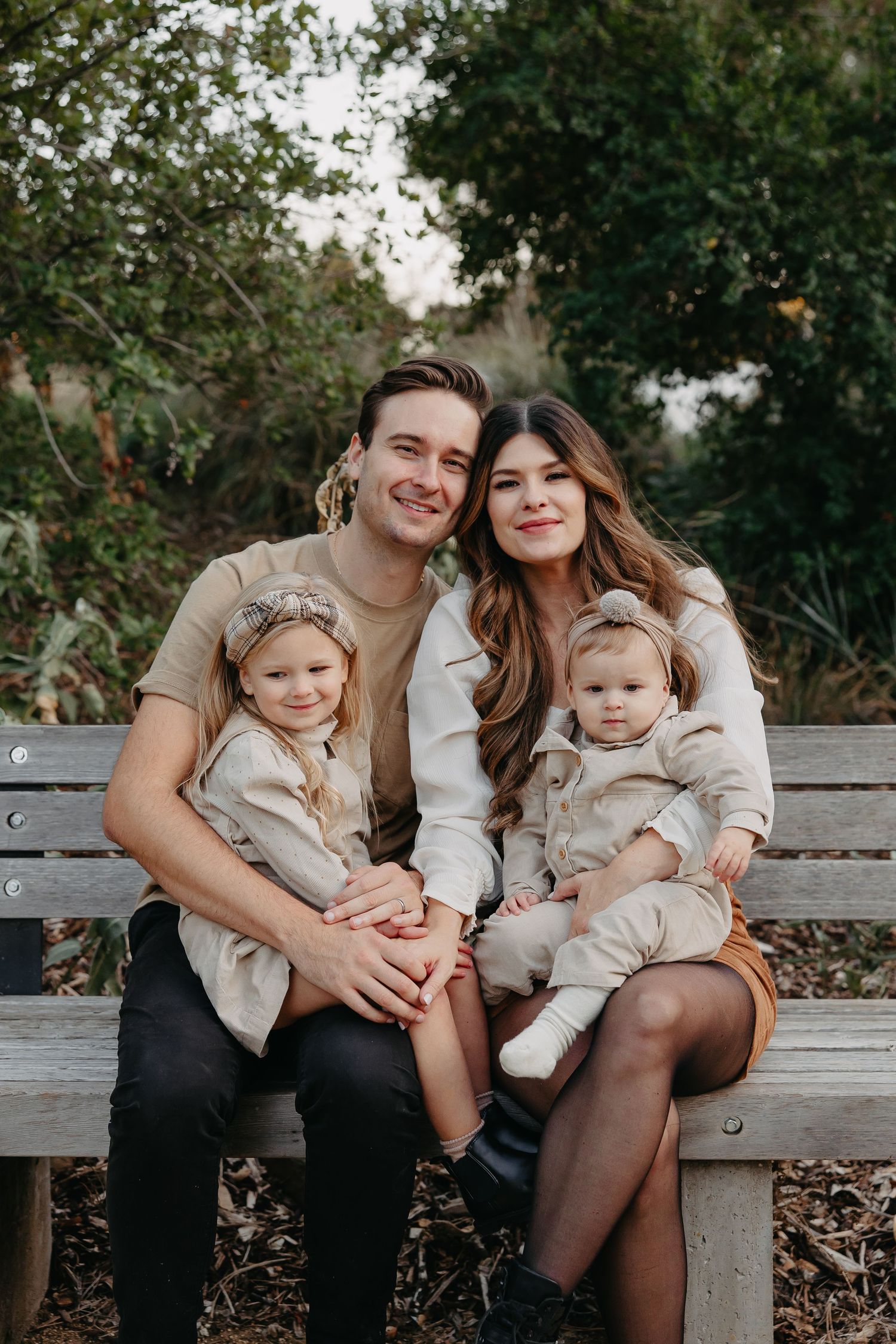 Happy family wearing coordinated beige and neutral outfits sitting together on wooden bench in outdoor setting.