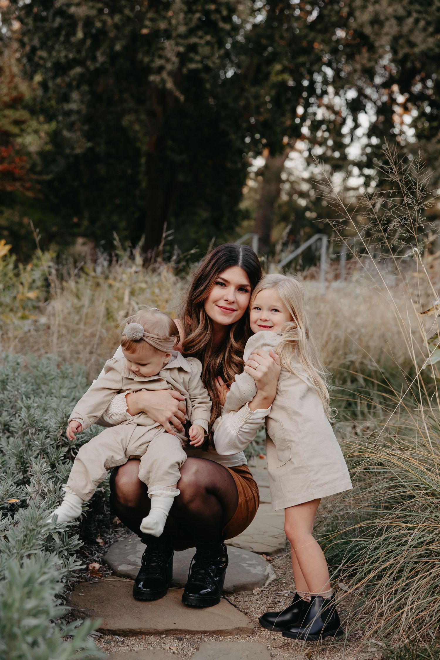 A family in coordinating neutral outfits poses outdoors among tall grasses and autumn foliage during golden hour.