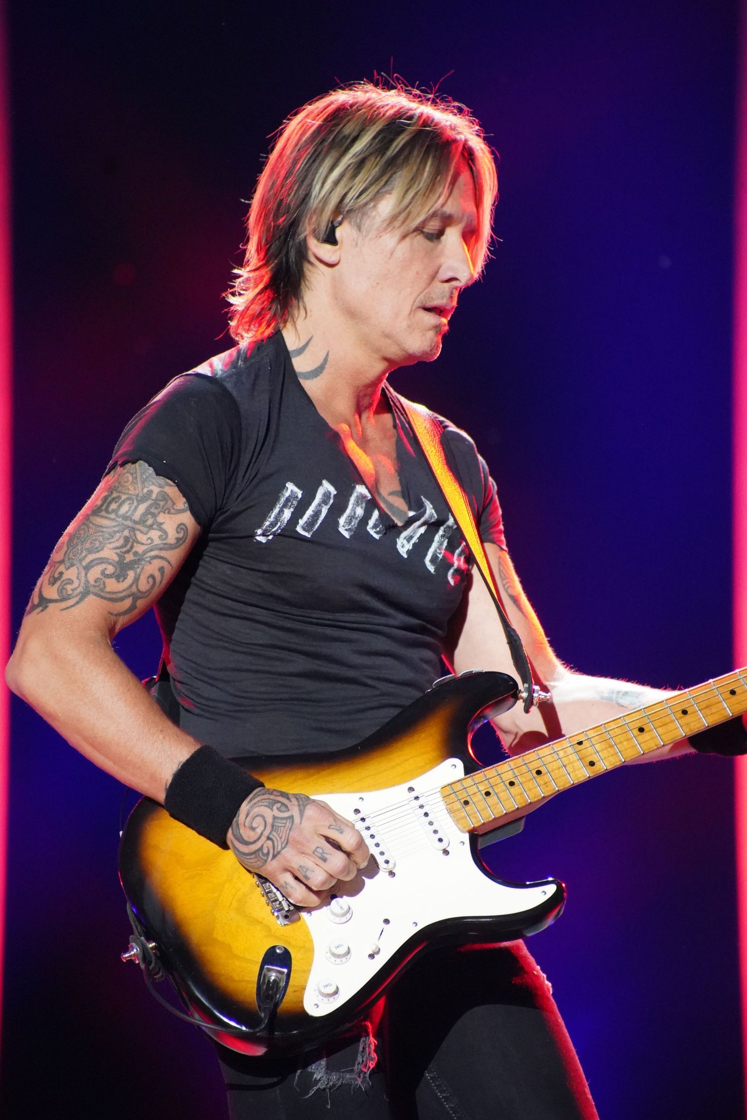 A musician in black t-shirt plays electric guitar during concert with dramatic lighting.