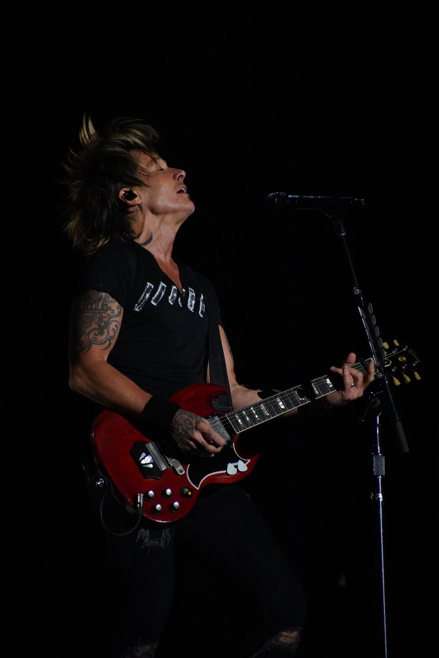 A musician plays red electric guitar while singing during a darkly lit performance.