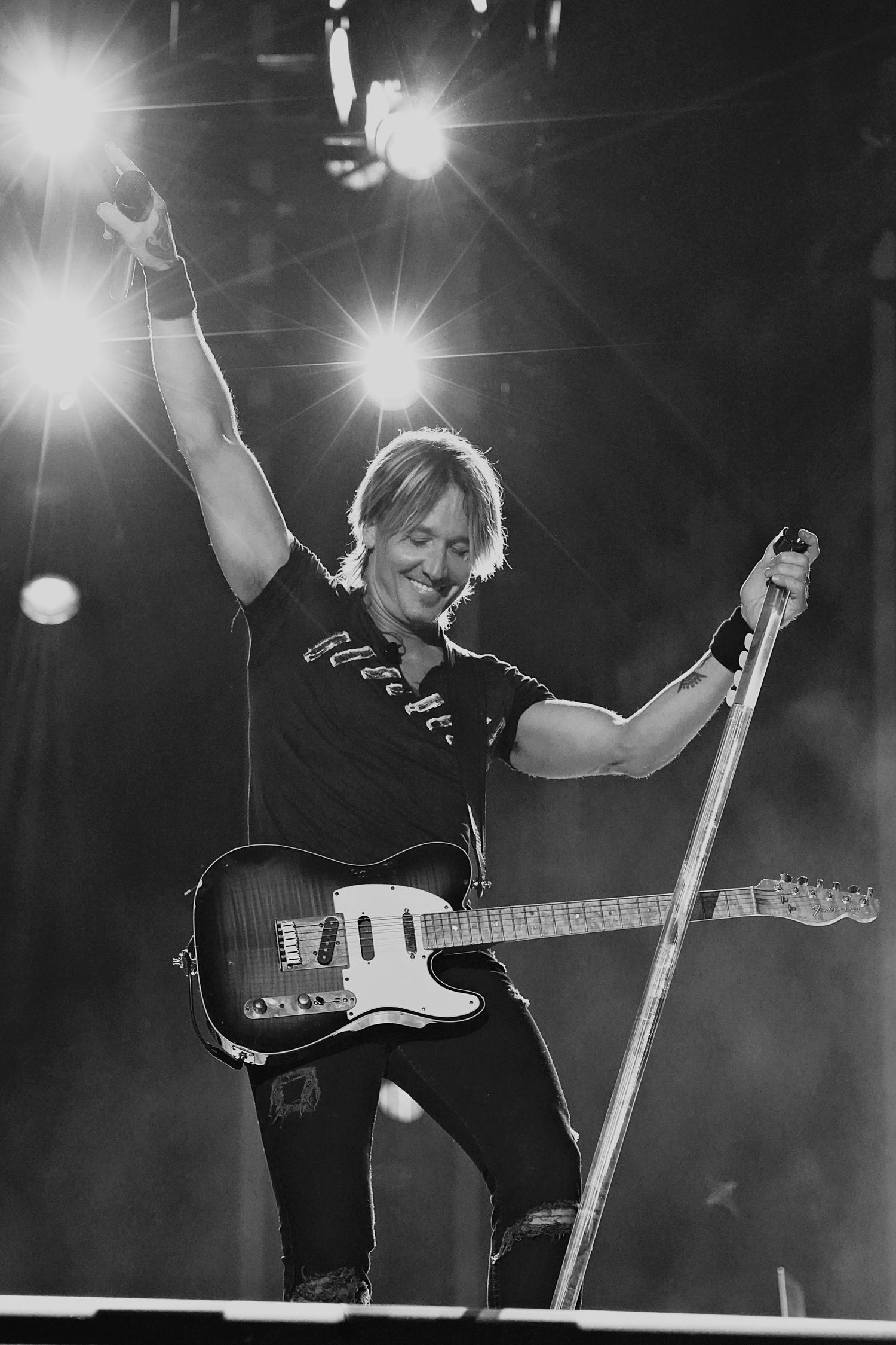 A performer in black t-shirt plays guitar and poses dramatically in black and white concert photo.
