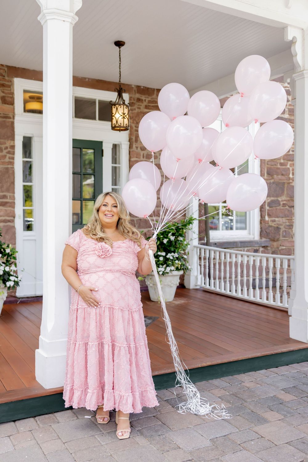 Pregnant woman in pink dress holding pink balloons on a porch at Orange Lawn Tennis Club.