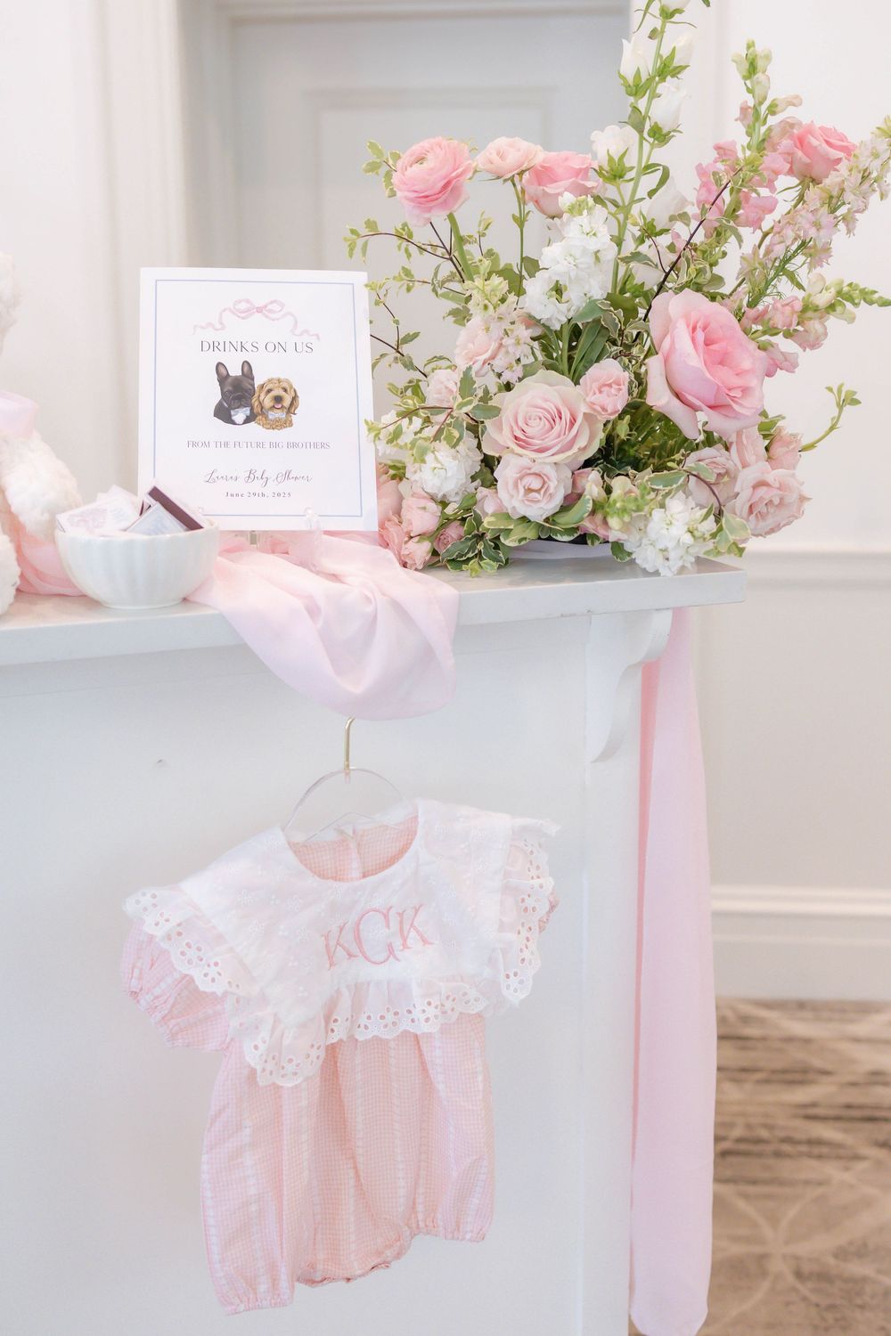 Pink baby shower display with monogrammed romper, floral arrangement, and welcome sign.