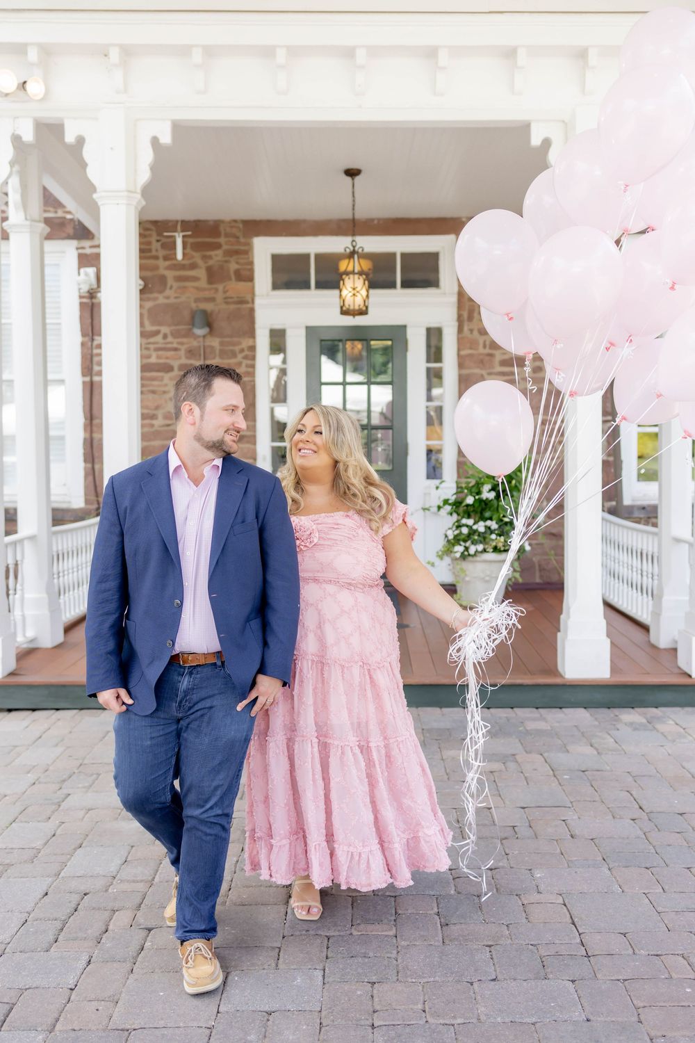 A couple walks together holding pink balloons outside Orange Lawn Tennis Club, wearing a navy suit and pink dress.