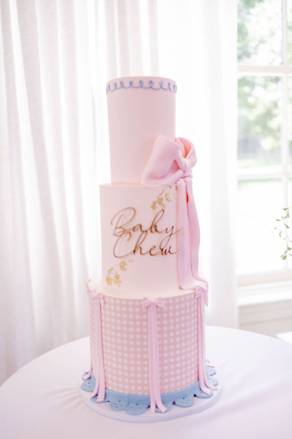 Three-tier pink baby shower cake with gingham pattern, bow, and gold script lettering.
