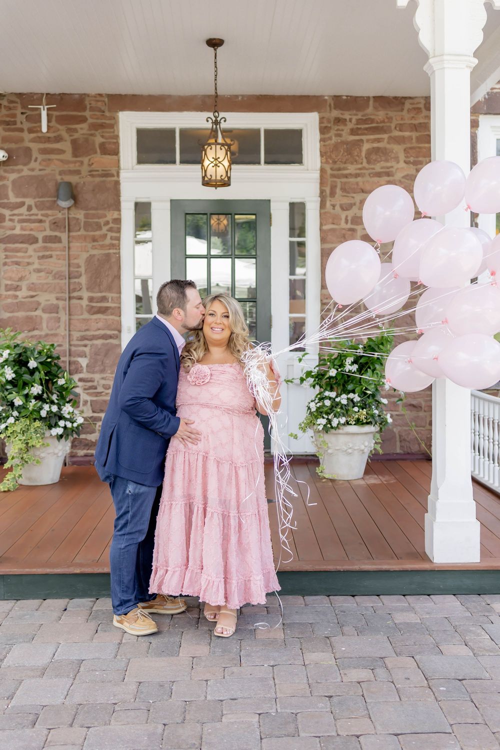 Couple poses on porch at Orange Lawn Tennis Club, man kissing woman in pink dress holding pink balloons at gender reveal.