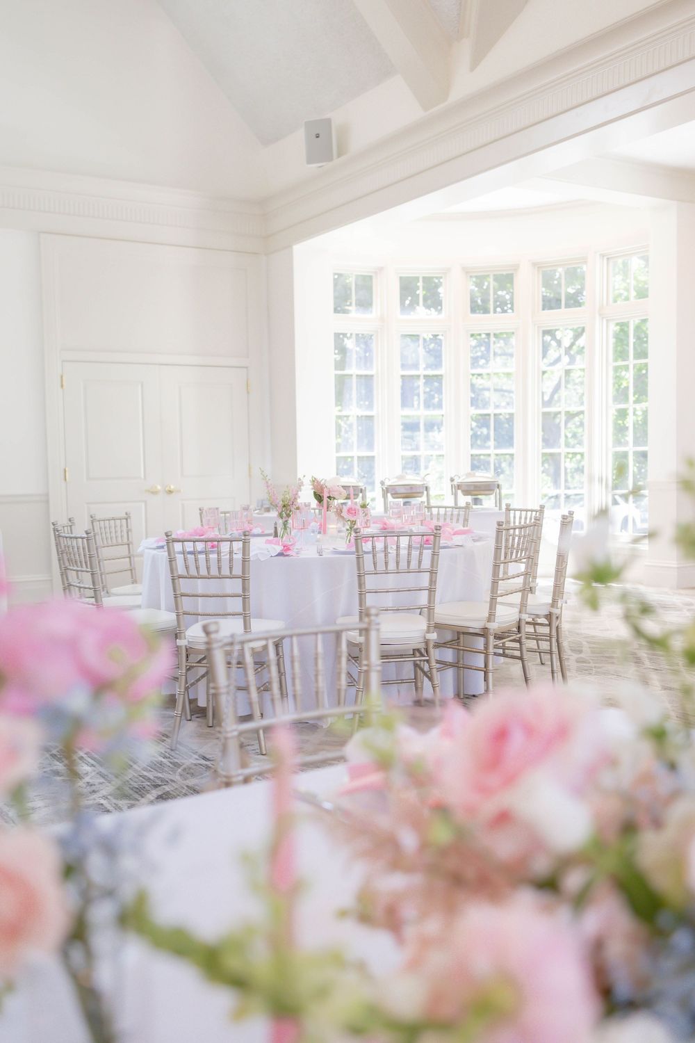 Elegant baby with white tables, gold chiavari chairs, and pink floral centerpieces in a bright, airy venue.