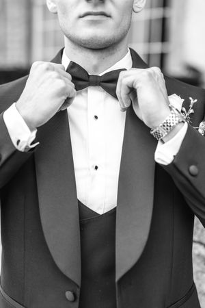Black and white photo of a groom in a tuxedo adjusting his bow tie, with a boutonniere and watch visible.