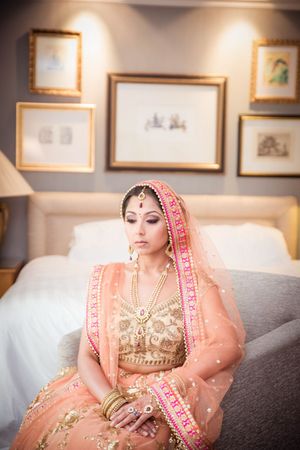 Indian bride in peach lehenga with gold embroidery and pink dupatta sits gracefully in elegant room.