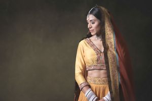 Young woman in traditional yellow Indian lehenga with red dupatta and gold jewelry poses gracefully in studio.