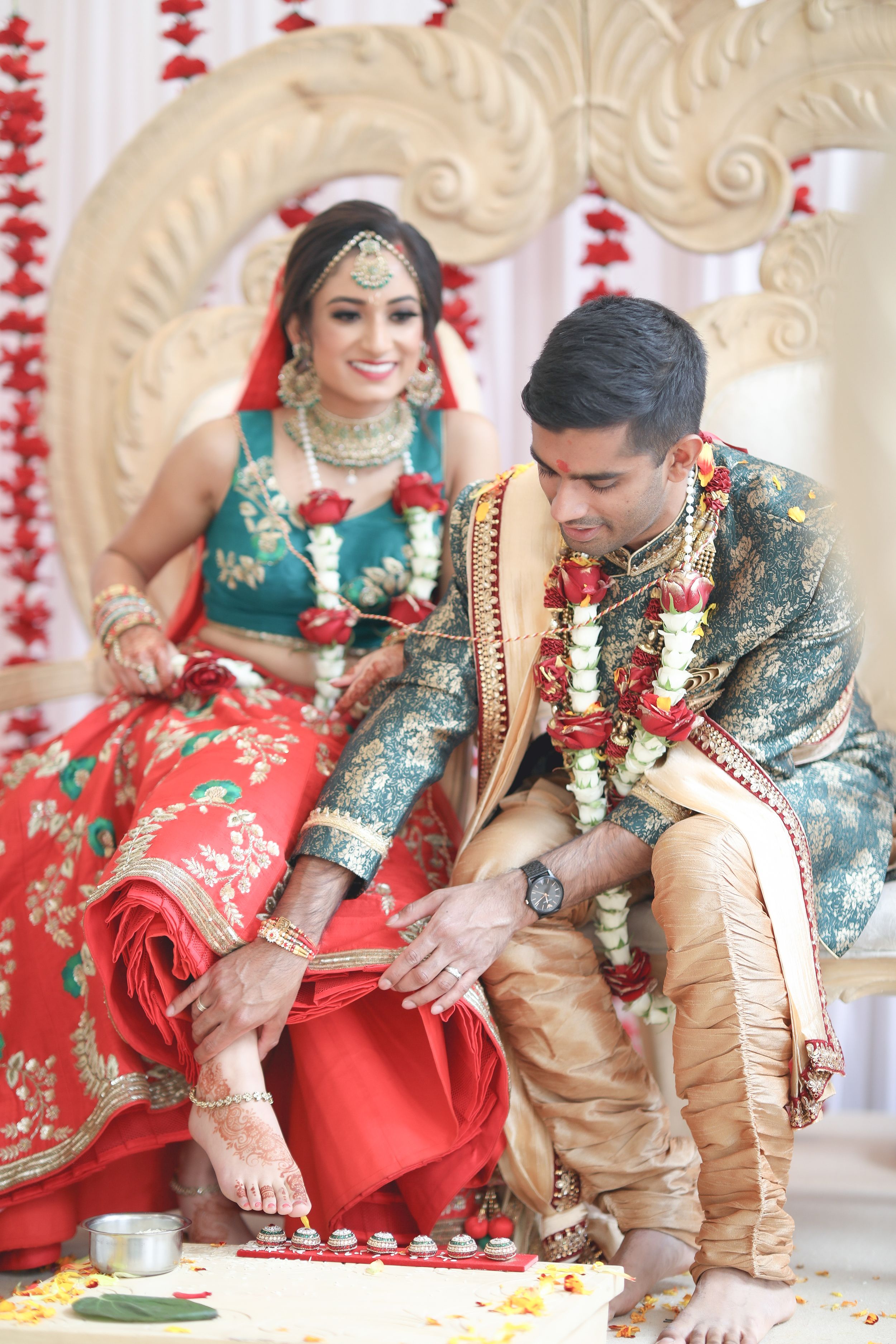 Indian bride in red lehenga and groom in teal sherwani performing traditional Hindu wedding rituals on decorated mandap.