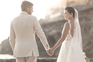 A bride and groom hold hands at sunset near rocky cliffs, she in a lace veil and he in a beige linen suit.