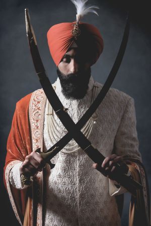 Sikh man in orange turban and traditional sherwani holding two crossed curved swords in dramatic studio portrait.