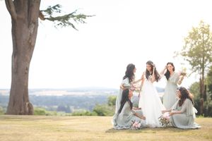 Bride in white gown surrounded by bridesmaids in grey dresses on a sunny lawn with a large tree backdrop.
