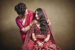 A mother helps her daughter in a red bridal lehenga with dupatta during an Indian wedding preparation.