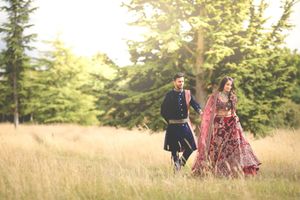 Couple in traditional South Asian wedding attire walking through golden meadow with lush green trees in background.