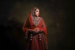 A bride in a red embroidered lehenga and dupatta poses gracefully in a dramatic studio portrait.