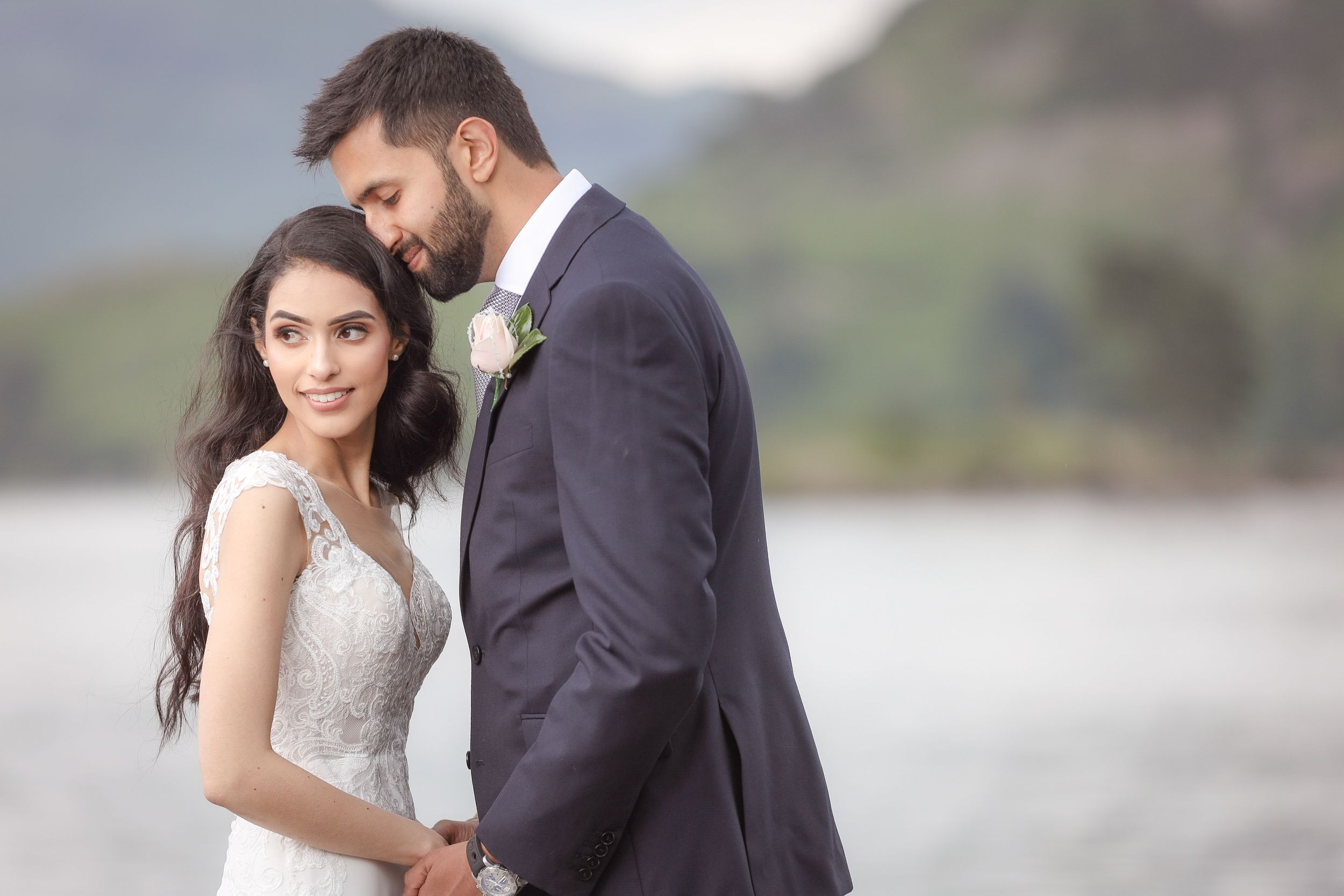 Elegant bride and groom posing outdoors near a lake with mountain backdrop on their wedding day.
