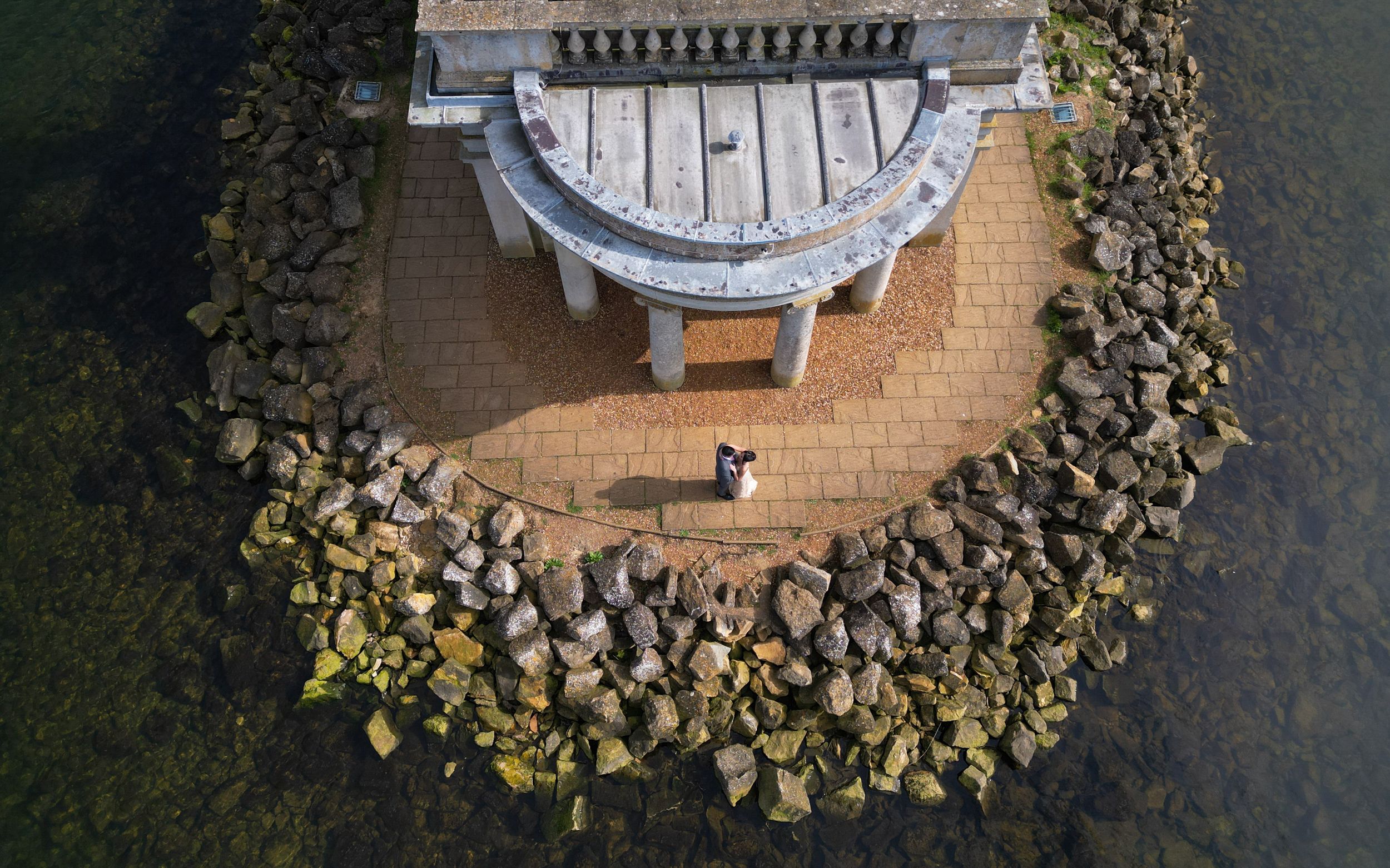 Aerial view of a couple standing on a brick pathway beside a stone gazebo surrounded by rocks and water.