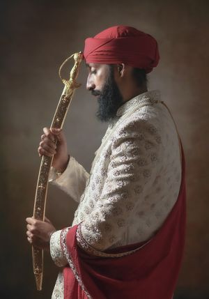 Bearded Sikh man in ornate white sherwani and red turban holding a decorative gold sword in a studio portrait.