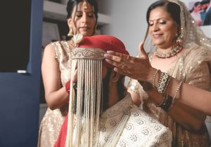 Two women in traditional Indian attire during a bridal preparation ceremony, adorning a red dupatta with gold fringe.