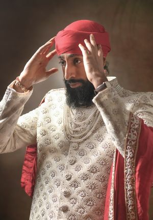 Indian groom in white embroidered sherwani adjusting red turban with pearl necklaces and traditional wedding attire.