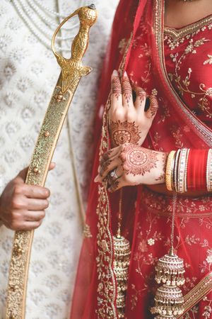 Indian bride in red lehenga with intricate mehndi henna hands and gold bangles beside groom holding ceremonial sword.