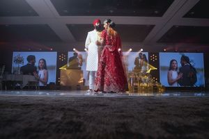 Indian bride and groom in traditional red and white wedding attire standing on stage at elegant banquet hall ceremony.