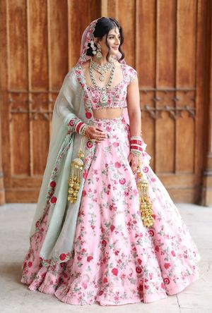 Bride in pink floral lehenga with mint dupatta and gold jewelry poses elegantly for Indian wedding portrait.