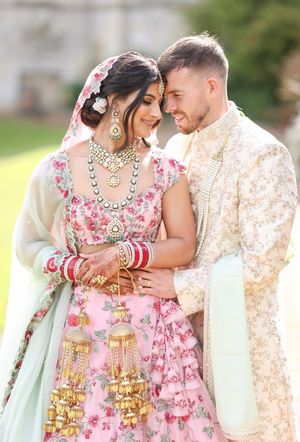 Couple in traditional Indian wedding attire, bride in floral pink lehenga with gold jewelry, groom in cream sherwani.