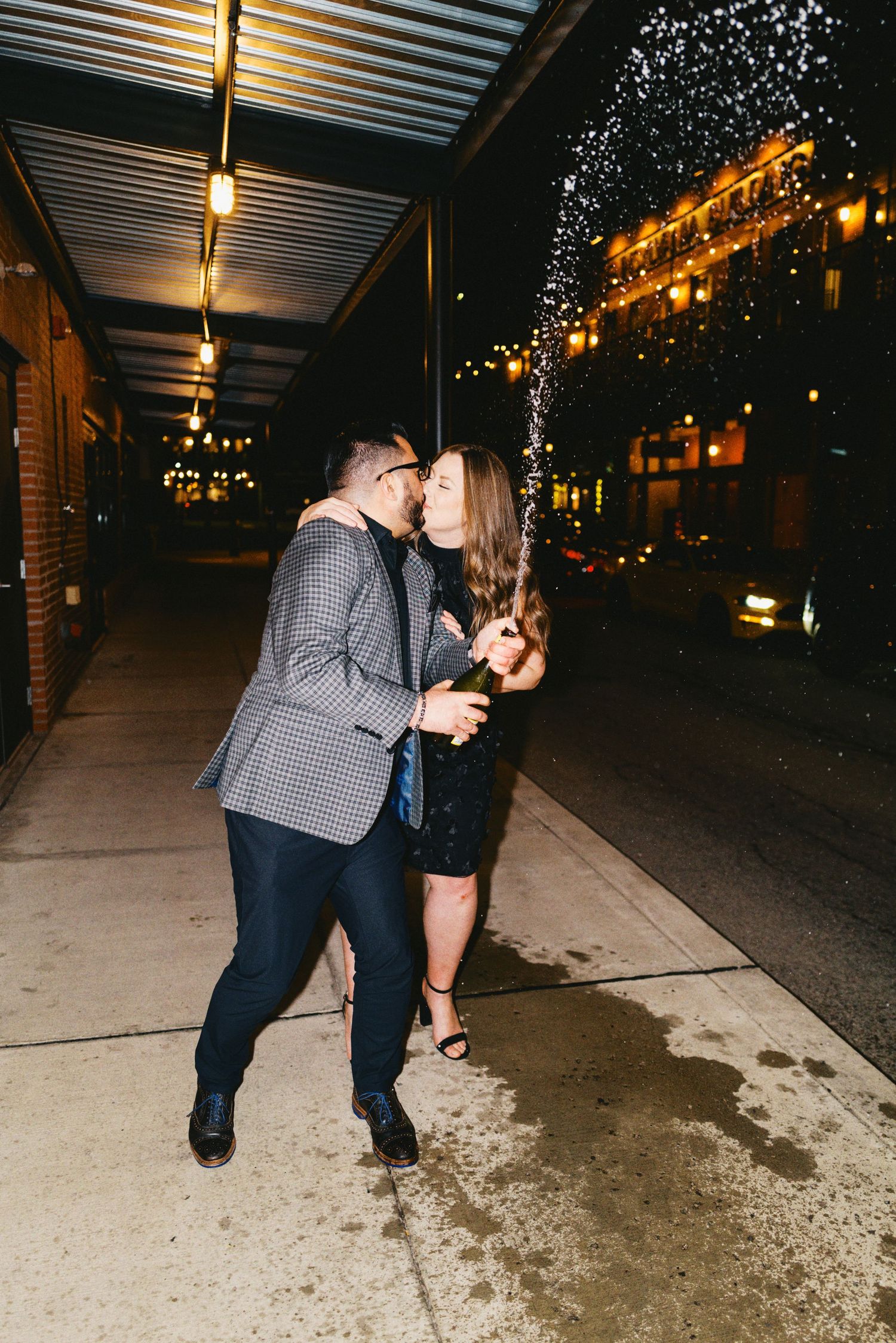 Couple kissing on a city sidewalk at night while popping champagne, dressed in elegant evening attire.