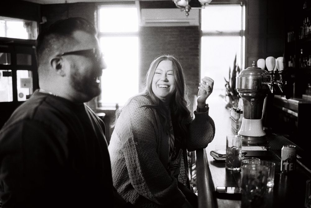 Two people laughing together at a bar counter in a candid black and white photo.