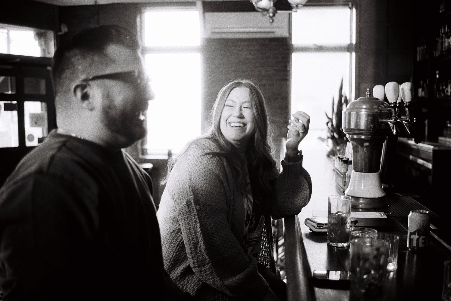 Two people laughing together at a bar counter in a candid black and white photo.