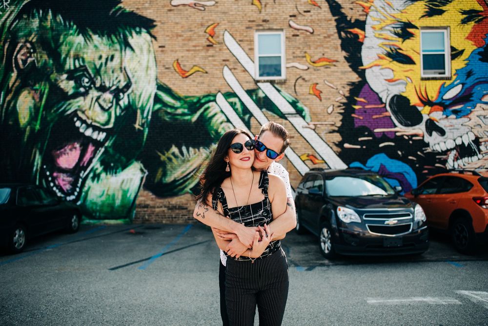 Couple embracing in front of vibrant street art murals featuring colorful graffiti characters on a brick wall.