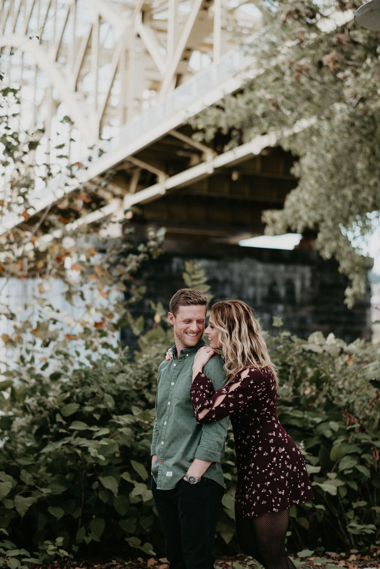 A couple shares a romantic moment under a bridge surrounded by lush greenery, the woman whispering in the mans ear.