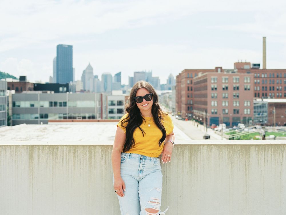 Young woman in yellow top and ripped jeans poses on rooftop with Pittsburgh city skyline in background.