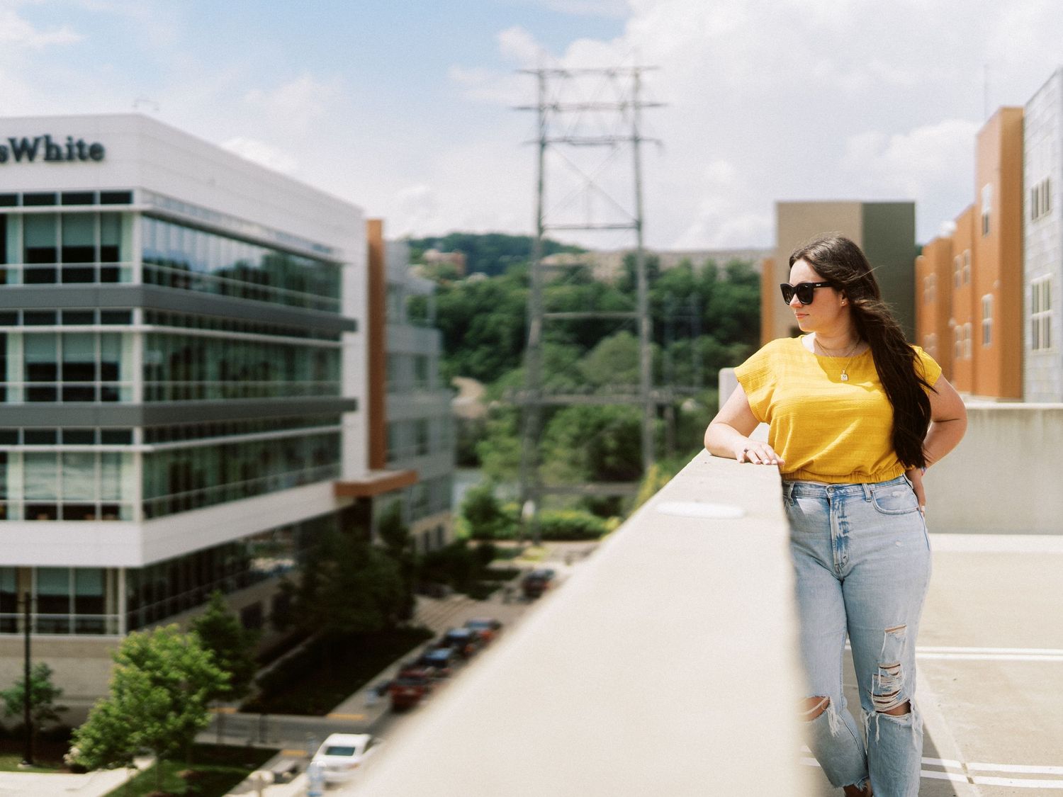 Woman in yellow top and ripped jeans leaning on rooftop railing with urban skyline background.