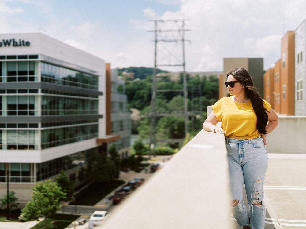 Woman in yellow top and ripped jeans leaning on rooftop railing with urban skyline background.