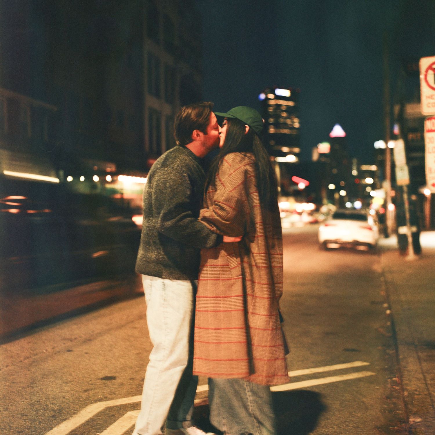 Couple sharing a romantic kiss on a city street at night, with glowing urban lights in the background.