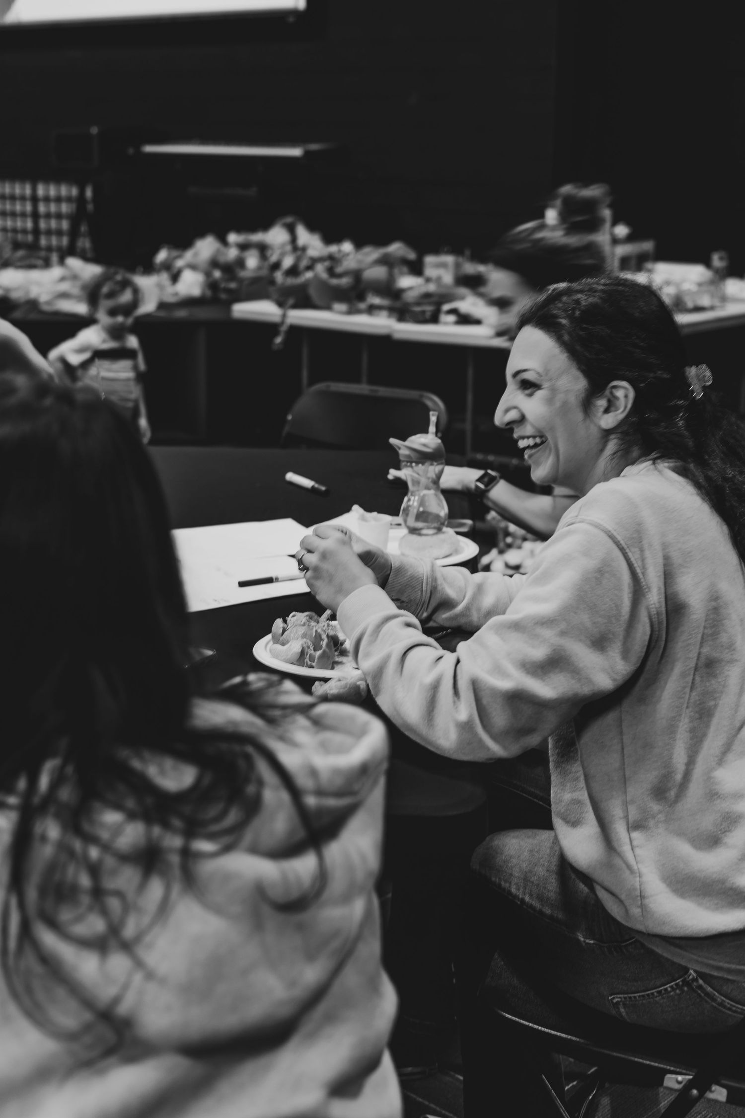 Black and white photo of a smiling woman eating at a table during a casual indoor gathering event.