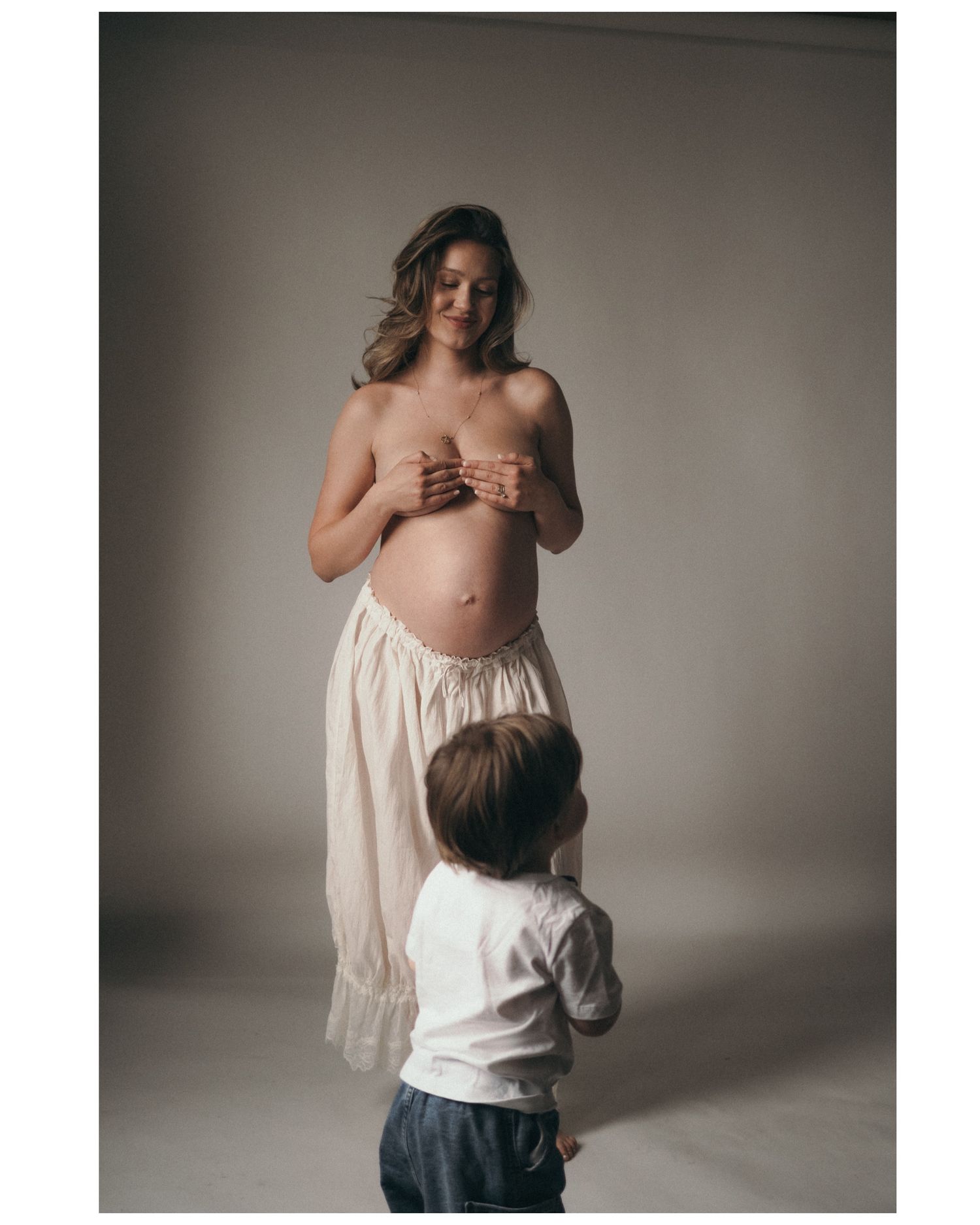 Pregnant woman in sheer skirt poses with young child in a soft-lit studio maternity photoshoot.