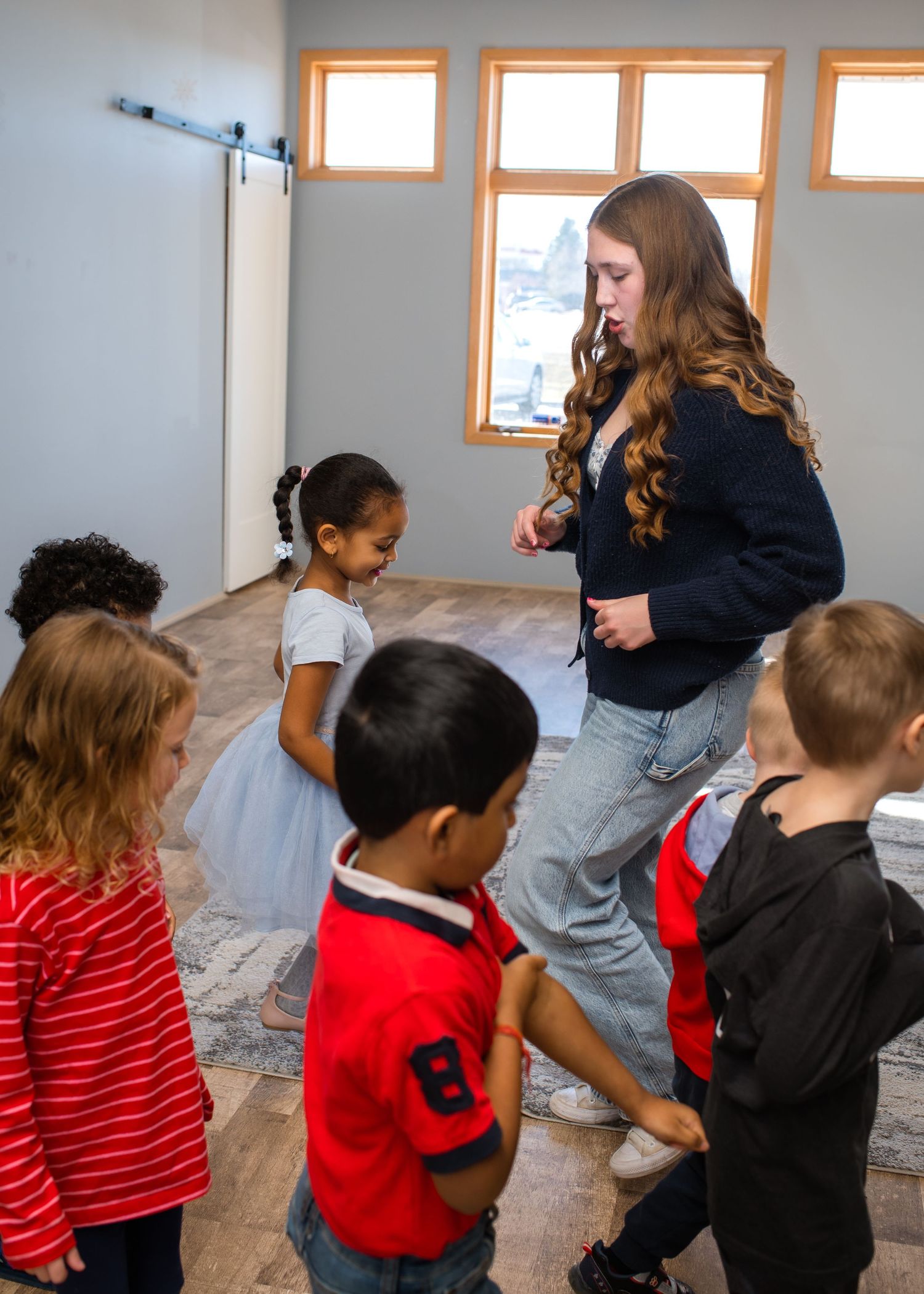 A young woman teacher interacts with a diverse group of small children in a bright modern classroom with barn door.
